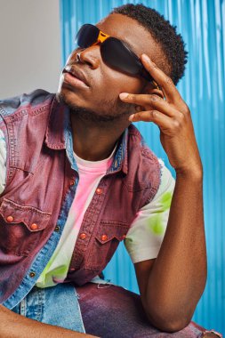 Güneş gözlüğü takmış şık Afroamerikan modelinin portresi. Renkli kot yelek ve tişört. Arka planda mavi polikarbonat çarşaf, moda çekimi, DIY kıyafetler, sürdürülebilir yaşam tarzı. 