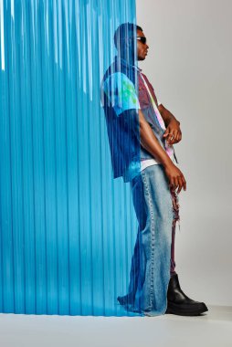 Gri arka planda mavi polikarbonat çarşafın arkasında duran yırtık kot pantolon, kot yelek ve güneş gözlüklü şık Afroamerikan erkek modelinin yan görünüşü, DIY giysileri, sürdürülebilir yaşam tarzı 