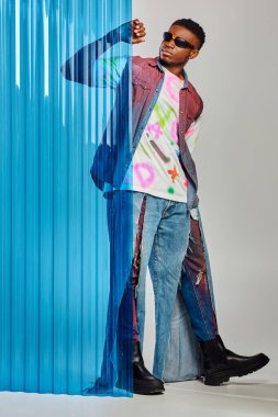 Güneş gözlüklü kot yelek ve yırtık kot pantolonlu yakışıklı Afroamerikan modeli mavi polikarbonat çarşafa dokunuyor ve gri arka planda duruyor, DIY giysileri, sürdürülebilir yaşam tarzı 