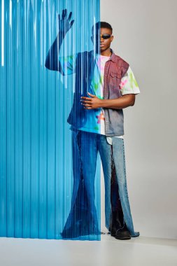 Güneş gözlüklü Afroamerikan bir adam, yırtık kot pantolon ve renkli kot yelek mavi polikarbonat çarşafın yanında poz veriyor ve gri arka plan, moda çekimi, DIY giysi, sürdürülebilir yaşam tarzı 
