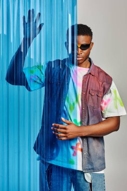 Trendy afroamerican male model in sunglasses, colorful t-shirt and denim vest posing behind blue polycarbonate sheet on grey background, fashion shoot, DIY clothing, sustainable lifestyle  clipart