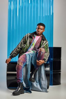 Full length of young and stylish afroamerican model in outwear jacket with led stripes and ripped jeans looking at camera while sitting near blue polycarbonate sheet on grey, DIY clothing  clipart
