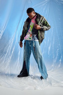 Üstü açık ceketli yakışıklı Afroamerikan bir adam kurşun çizgili modaya uygun yırtık pantolonlara bakıyor mavi arka planda parlak selofan üzerinde duruyor, şehir kıyafeti, DIY kıyafeti, sürdürülebilir yaşam tarzı 
