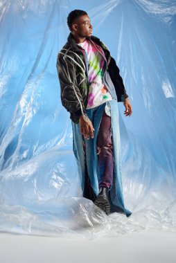 Üstü açık ceketli, mavi arka planda parlak selofan üzerinde yürüyen yırtık kot pantolonlu şık afroamerikan bir adam. Şehir kıyafeti ve modern poz, DIY giysi, sürdürülebilir yaşam tarzı. 