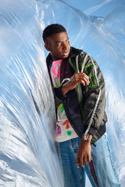 Trendy young afroamerican male model in ripped jeans touching outwear jacket with led stripes near glossy cellophane on blue background, urban outfit, DIY clothing, sustainable lifestyle  clipart