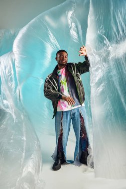 Full length of trendy young african american man in ripped jeans and outwear jacket looking away near glossy cellophane on turquoise background, urban outfit, creative expression, DIY clothing  clipart