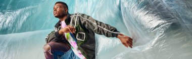 Modern ceketli ve yırtık pantolonlu genç Afroamerikalı adam turkuaz arka planda selofan pantolonu, kentsel kıyafet ve modern poz, afiş, yaratıcı ifade, DIY giysi 