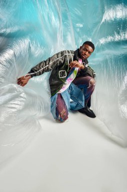 Fashionable and confident young african american model in outwear jacket and ripped jeans looking at camera near cellophane on turquoise background, creative expression, DIY clothing  clipart