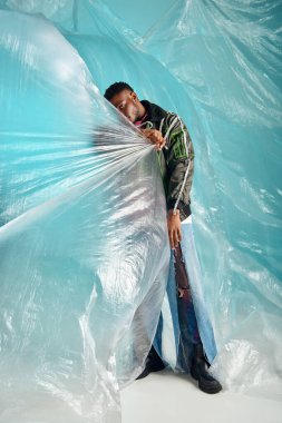 Yırtık kot pantolon ve uzun ceket giyen, turkuaz arka planda selofan ile yüzünü örten Afro-Amerikan erkeği, kentsel kıyafet ve modern poz, yaratıcı ifade, DIY giysi 