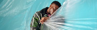 Young and trendy afroamerican model in outwear jacket and led stripes covering face with cellophane on turquoise background, urban outfit and modern pose, banner, creative expression, DIY clothing  clipart
