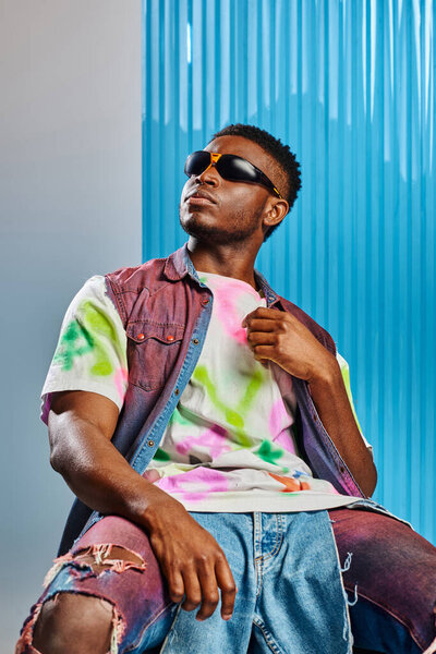 Portrait of stylish young afroamerican man in sunglasses, colorful t-shirt and denim vest on grey with blue polycarbonate sheet at background, fashion shoot, sustainable fashion, DIY clothing