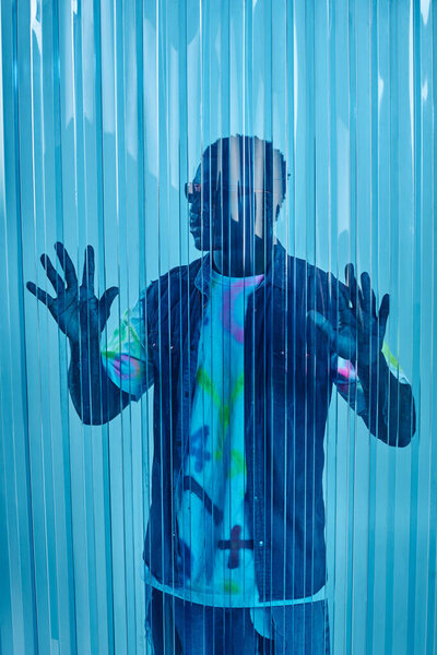 Stylish afroamerican man in sunglasses, denim vest and colorful t-shirt touching blue polycarbonate sheet, fashion shoot and street style outfit, DIY clothing, sustainable lifestyle 