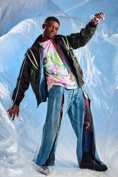 Full length of confident afroamerican man in outwear jacket with led stripes and ripped jeans looking at camera near glossy cellophane on blue background, modern pose, sustainable lifestyle 