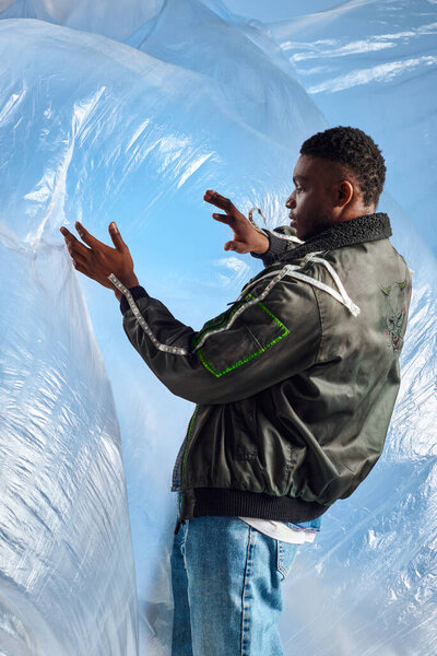 Side view of stylish african american man in jacket with led stripes and ripped jeans touching glossy cellophane on blue background, urban outfit and modern pose, creative expression, DIY clothing 