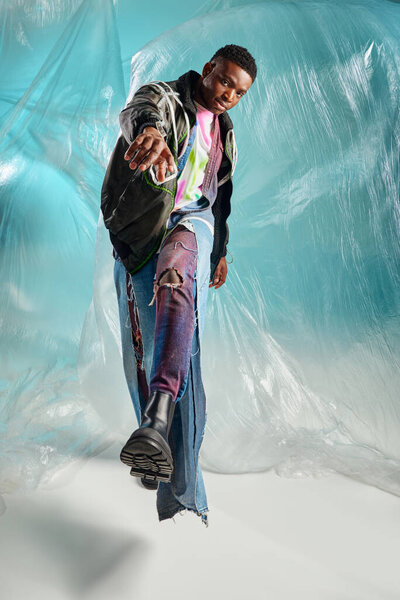 Full length of trendy young afroamerican model in outwear jacket and ripped jeans looking at camera and walking near glossy cellophane on turquoise background, creative expression, DIY clothing 