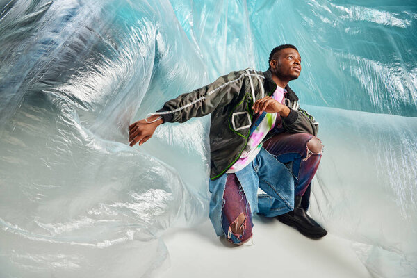 Young afroamerican model in outwear jacket with led stripes and ripped jeans looking away near cellophane on turquoise background, urban outfit and modern pose, creative expression, DIY clothing 