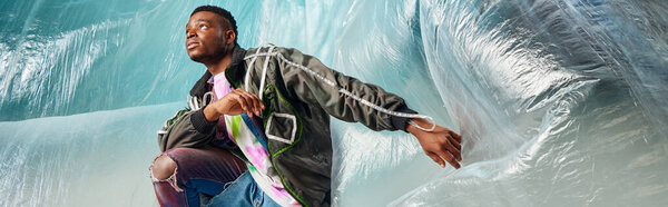 Young afroamerican man in modern outwear jacket and ripped jeans looking away near cellophane on turquoise background, urban outfit and modern pose, banner, creative expression, DIY clothing 