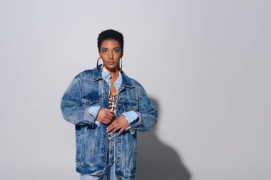 Fashionable african american woman with short hair looking at camera while posing in denim jacket and golden earrings on grey background, denim fashion concept clipart
