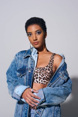 Portrait of fashionable and confident african american woman with bold makeup posing in top with animal print and denim jacket on grey background, denim fashion concept clipart