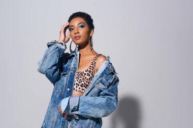 Confident young african american model with bold makeup touching short hair while posing in denim jacket and top with animal print on grey background, denim fashion concept clipart