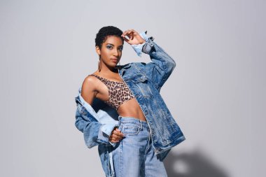Confident young african american woman with short hair posing in top with animal print, jeans and denim jacket while standing on grey background, denim fashion concept clipart