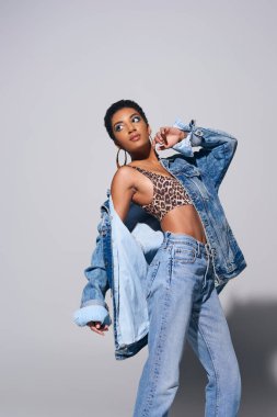 Kısa saçlı, canlı makyajlı, altın küpeli, kot ceketli, bluzlu ve kot pantolonlu, gri arka planda duran, kot pantolon giyen modacı Afro-Amerikan modeli.