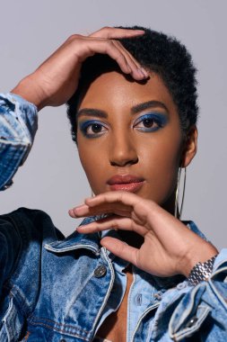 Portrait of modern african american woman with short hair and bold makeup touching face and looking at camera while posing in denim outfit isolated on grey, denim fashion concept clipart