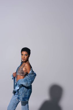 Fashionable african american woman with golden earrings and short hair posing in top with animal print and denim jacket on grey background, denim fashion concept clipart