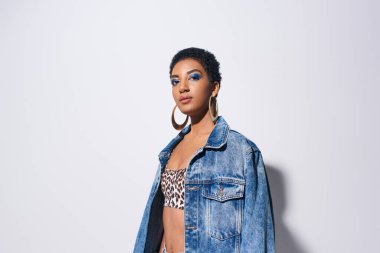 Portrait of confident african american model with bold makeup and golden earrings looking at camera while posing in top with animal print and denim jacket on grey background, denim fashion concept clipart