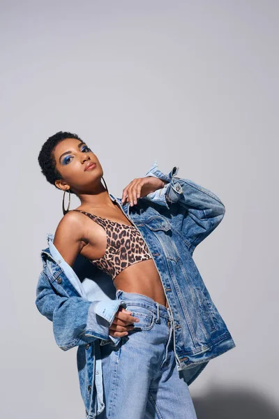 stock image Modern african american woman with bold makeup posing in top with animal print, denim jacket and jeans while standing on grey background, denim fashion concept
