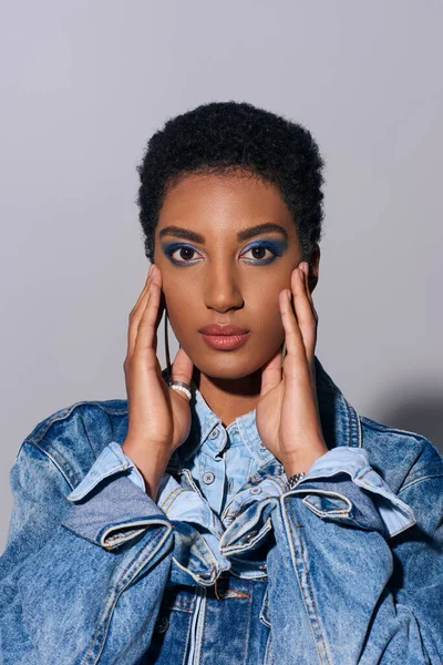 Young african american model with vivid makeup and short hair touching cheeks and posing in denim clothes while standing on grey background, denim fashion concept