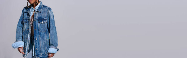 Обрезанный вид на стильную африканскую американскую женщину с золотыми серьгами, позирующими в верхней части с животным принтом и джинсовой курткой, стоя изолированы на сером, джинсовой концепции моды, баннер 