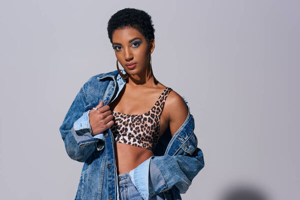 Confident young african american woman with golden earrings and vivid makeup posing in denim clothes and jeans while standing on grey background, denim fashion concept