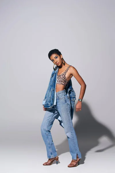 Trendy african amercan woman in top with animal print, denim jacket and blue jeans looking at camera while posing and standing on grey background, denim fashion concept