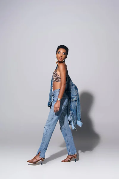 Full length of modern african american woman with golden earrings wearing top with animal print, denim jacket and jeans while walking on grey background, denim fashion concept