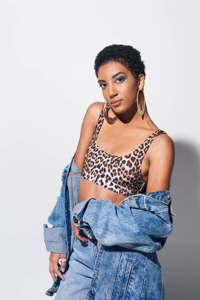 Stylish african american woman with bold makeup and short hair posing in top with animal print and casual denim attire while standing on grey background, denim fashion concept