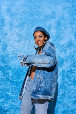 Canlı makyajlı, bereli, kot pantolonlu ve kot ceketli neşeli Afro-Amerikan modeli mavi desenli arka planda poz veriyor.