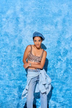 Bereli modaya uygun genç Afro-Amerikan kadın, kameraya gülümserken leopar desenli ve kot pantolonlu, mavi desenli arka planda duran, şık kot giyimli