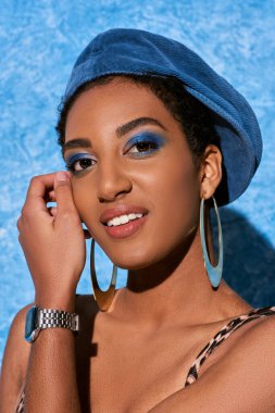 portrait of young african american woman with bold makeup, beret and golden earrings smiling at camera and posing on blue textured background, stylish denim attire clipart