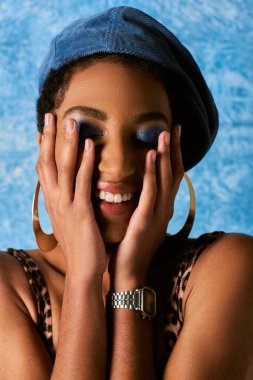 Portrait of positive african american woman with vivid makeup and beret touching cheeks and closing eyes on blue textured background, stylish denim attire clipart