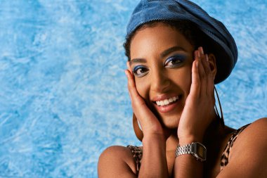 Portrait of trendy young african american model with bold makeup, earrings and beret touching cheeks and smiling at camera on blue textured background, stylish denim attire clipart