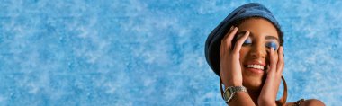 Trendy african american woman with bold makeup and denim beret smiling and touching face on blue textured background with copy space, stylish denim attire, banner  clipart