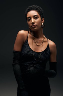 Siyah gece elbisesi, eldivenleri ve altın aksesuarlarıyla siyah, yüksek moda ve akşam kıyafetleri içinde şık ve modaya uygun bir şekilde kameraya bakan Afro-Amerikan kadın.