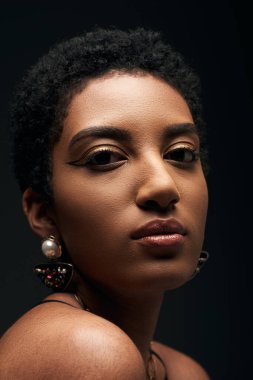 Genç ve kısa saçlı siyah saçlı, küpeli ve akşam makyajlı Afro-Amerikan kadını yakın çekimde siyah ışıkla izole edilmiş kameraya bakıyor.