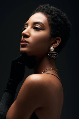 Şık ve kısa saçlı Afro-Amerikan kadın, gece makyajı ve altın aksesuarları ile, siyah üzerinde aydınlık, yüksek moda ve akşam görünümü ile tek başına dikilirken başka tarafa bakıyor.
