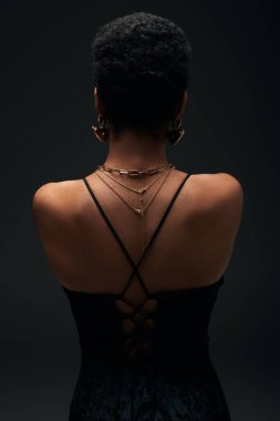 Back view of fashionable african american woman with short hair and golden accessories standing isolated on black with lighting, high fashion and evening look, jewelry, feminine, sensuality  clipart