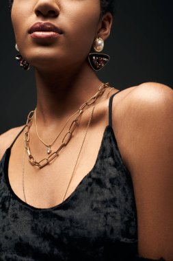 Cropped view of young and elegant african american model in dress and golden chains and earrings standing isolated on black under lighting, high fashion and evening look, details, close up clipart