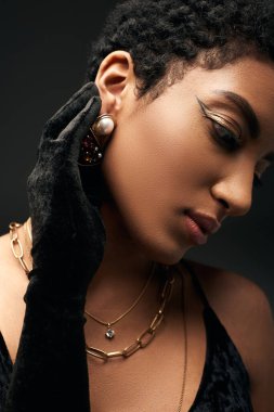 Akşam makyajlı, altın kolyeli ve siyah, yüksek moda ve akşam kıyafetlerine bürünmüş dokunaklı küpeli Afro-Amerikan kadın portresi.