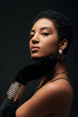 Akşam makyajı, eldiven ve altın aksesuarları olan özgüvenli ve zarif Afro-Amerikan kadın portresi siyah, yüksek moda ve akşam modasında izole edilmiş kameraya bakıyor.