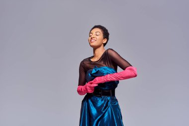 Cheerful and stylish young african american woman in pink gloves and cocktail dress standing with closed eyes isolated on grey, modern generation z fashion concept clipart